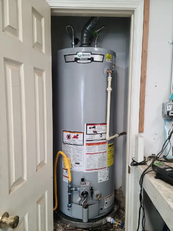 Tall gray water heater tank installed in a small closet with pipes and warning labels on it.