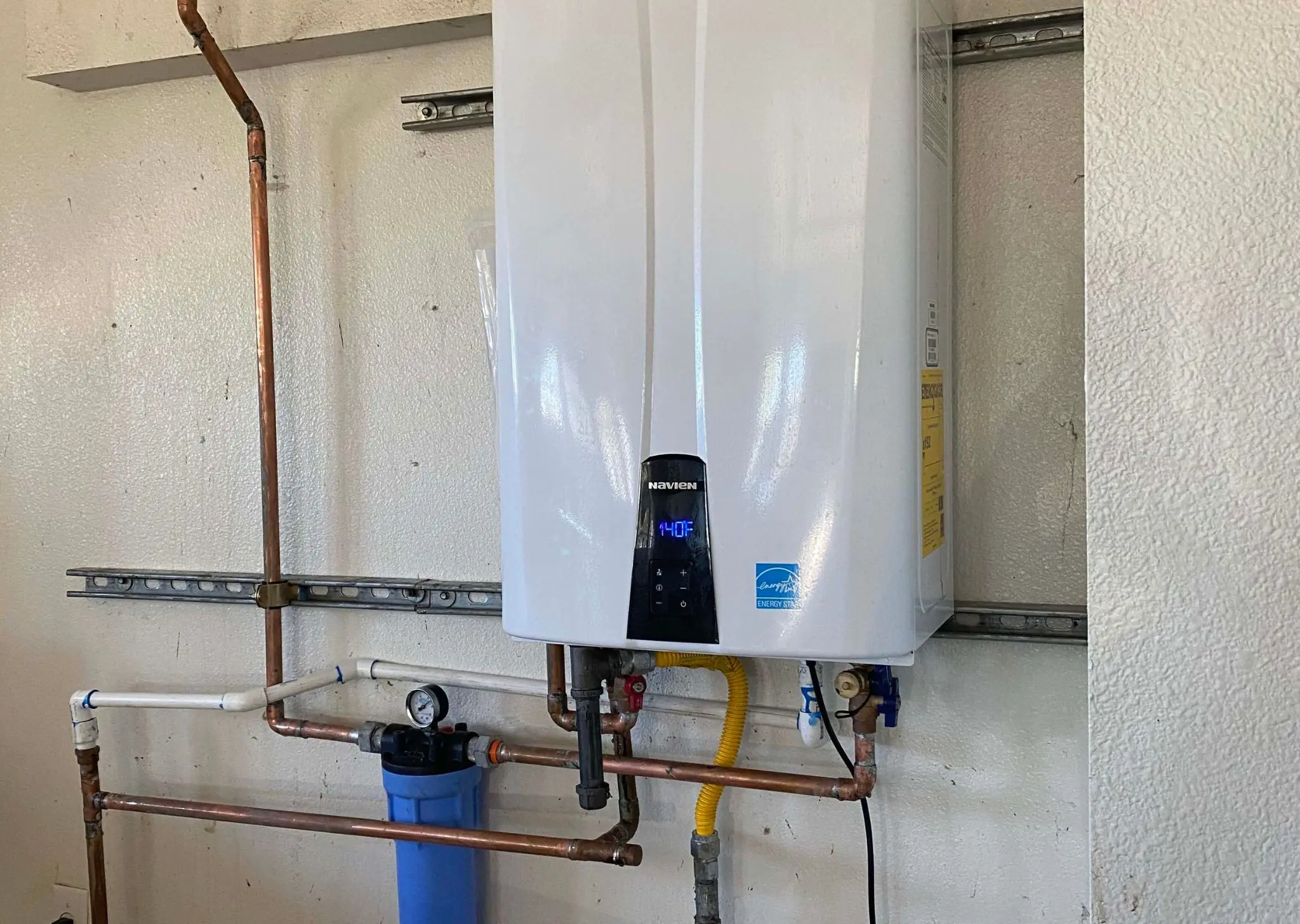 Navien tankless water heater mounted on wall with connected copper pipes and blue water filter.