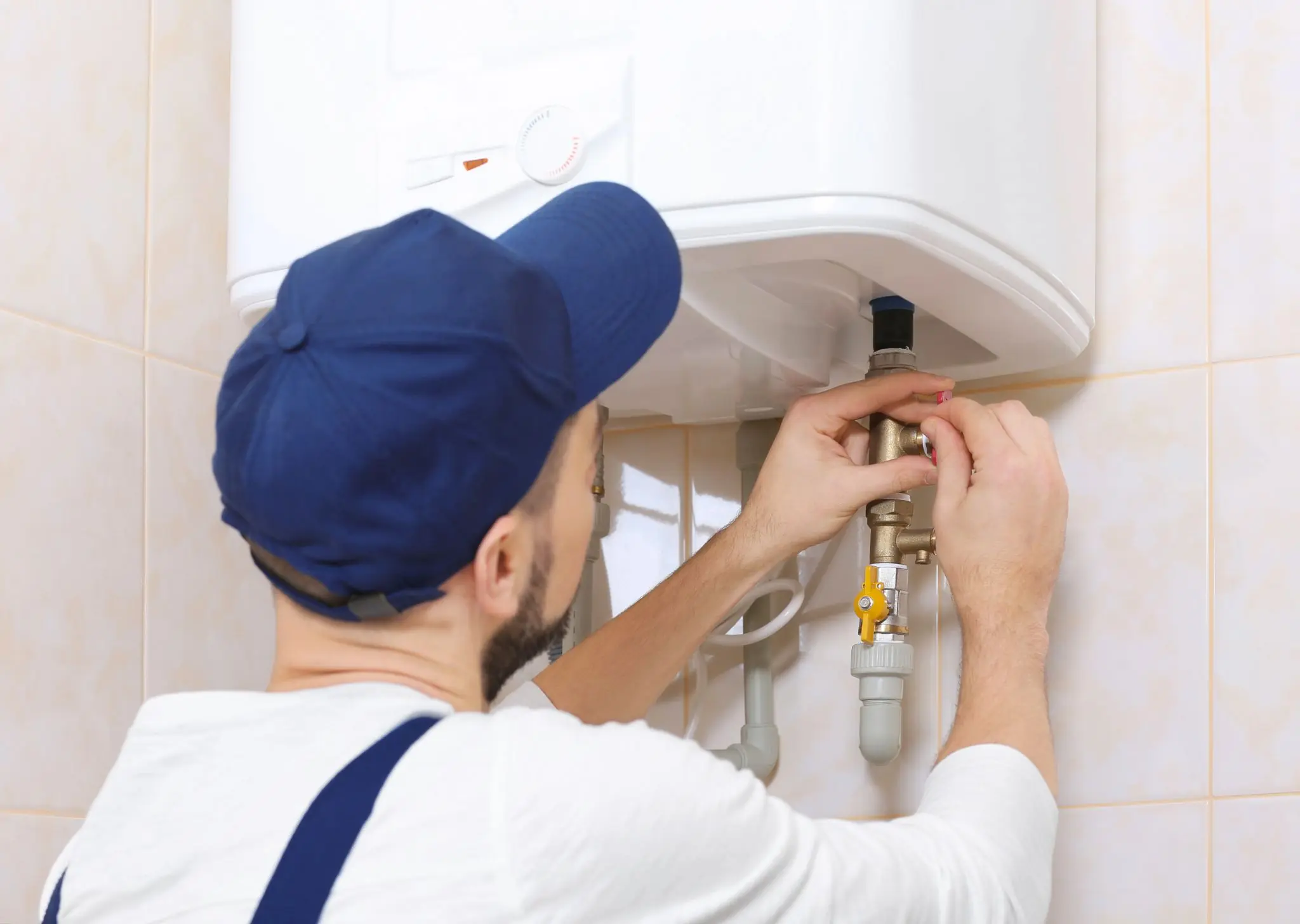 Technician in a blue cap installing or adjusting a tankless water heater on a tiled wall.