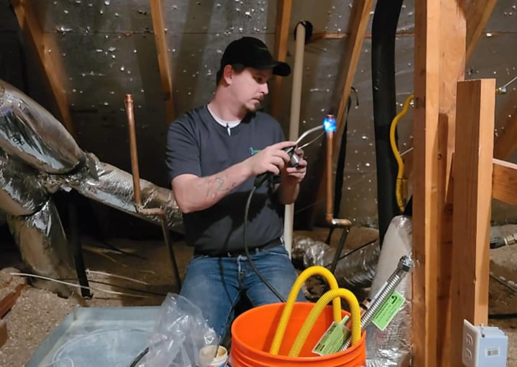 Worker using a torch to solder copper pipes in an attic with ducts and wooden beams.