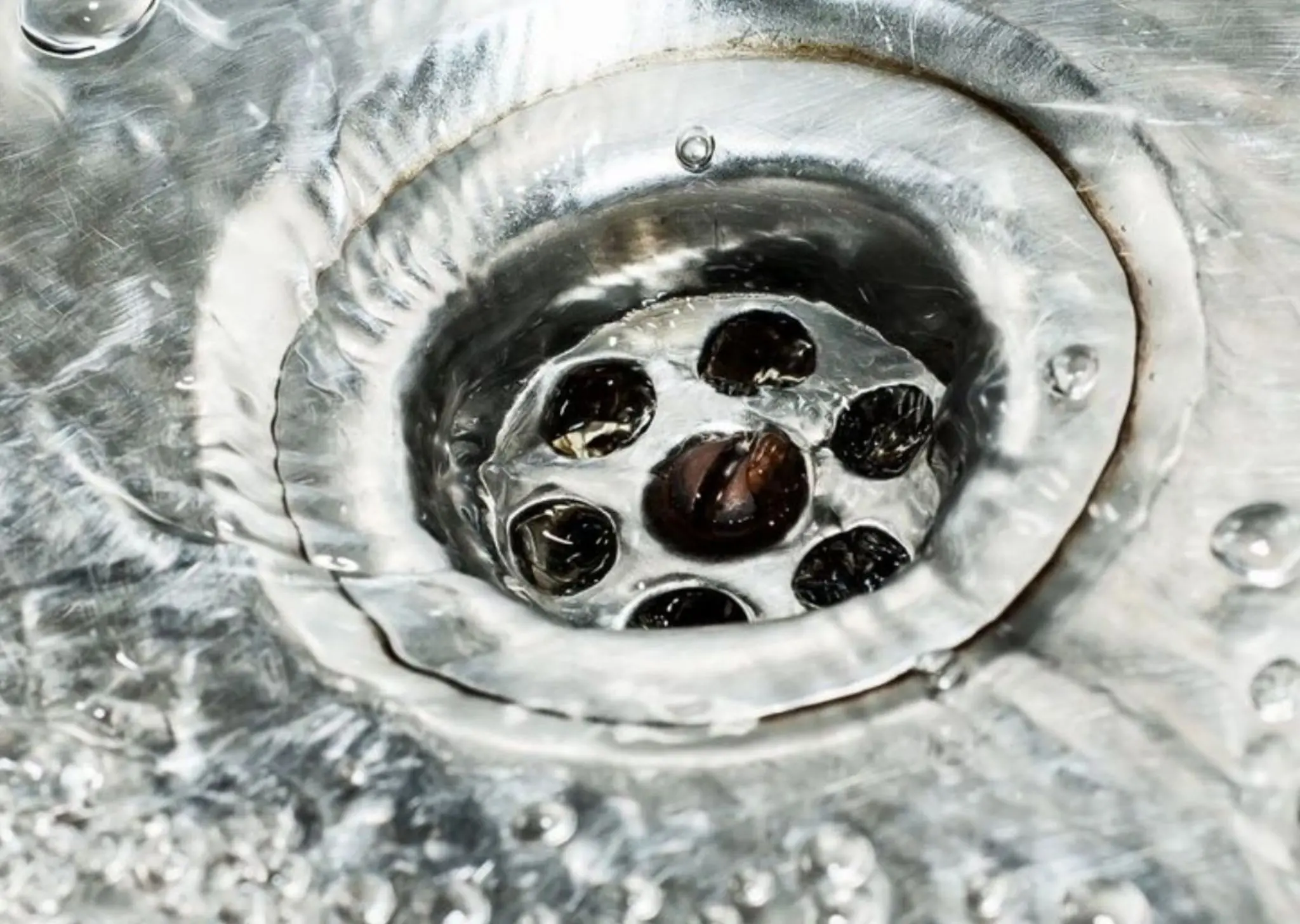 Close-up of water swirling down a stainless steel sink drain with bubbles on the surface.