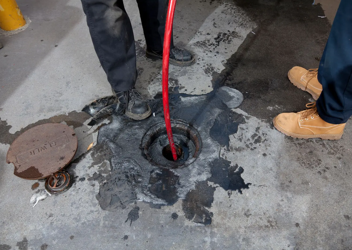 Worker using a red hose to clean a sewer drain with the manhole cover removed.