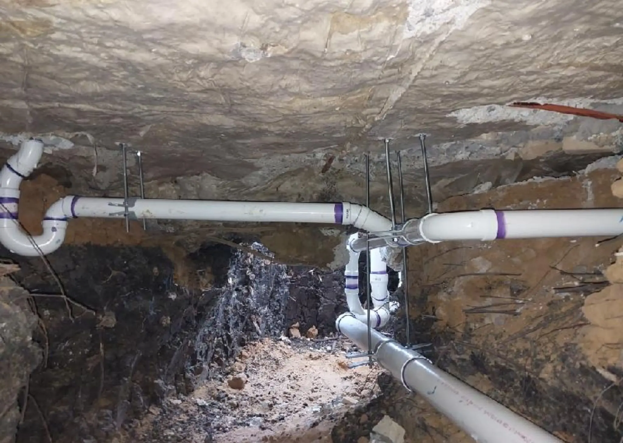 White PVC pipes with purple primer installed underground in a dirt trench supported by metal brackets.