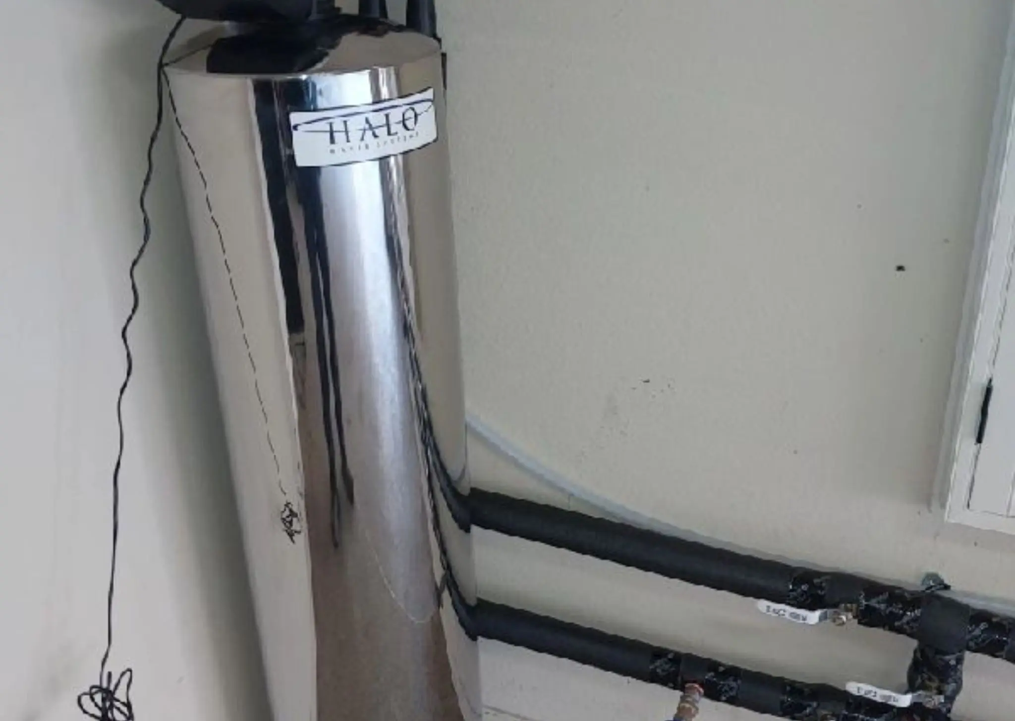 Silver HALO water treatment system unit connected to black insulated pipes inside a utility area.