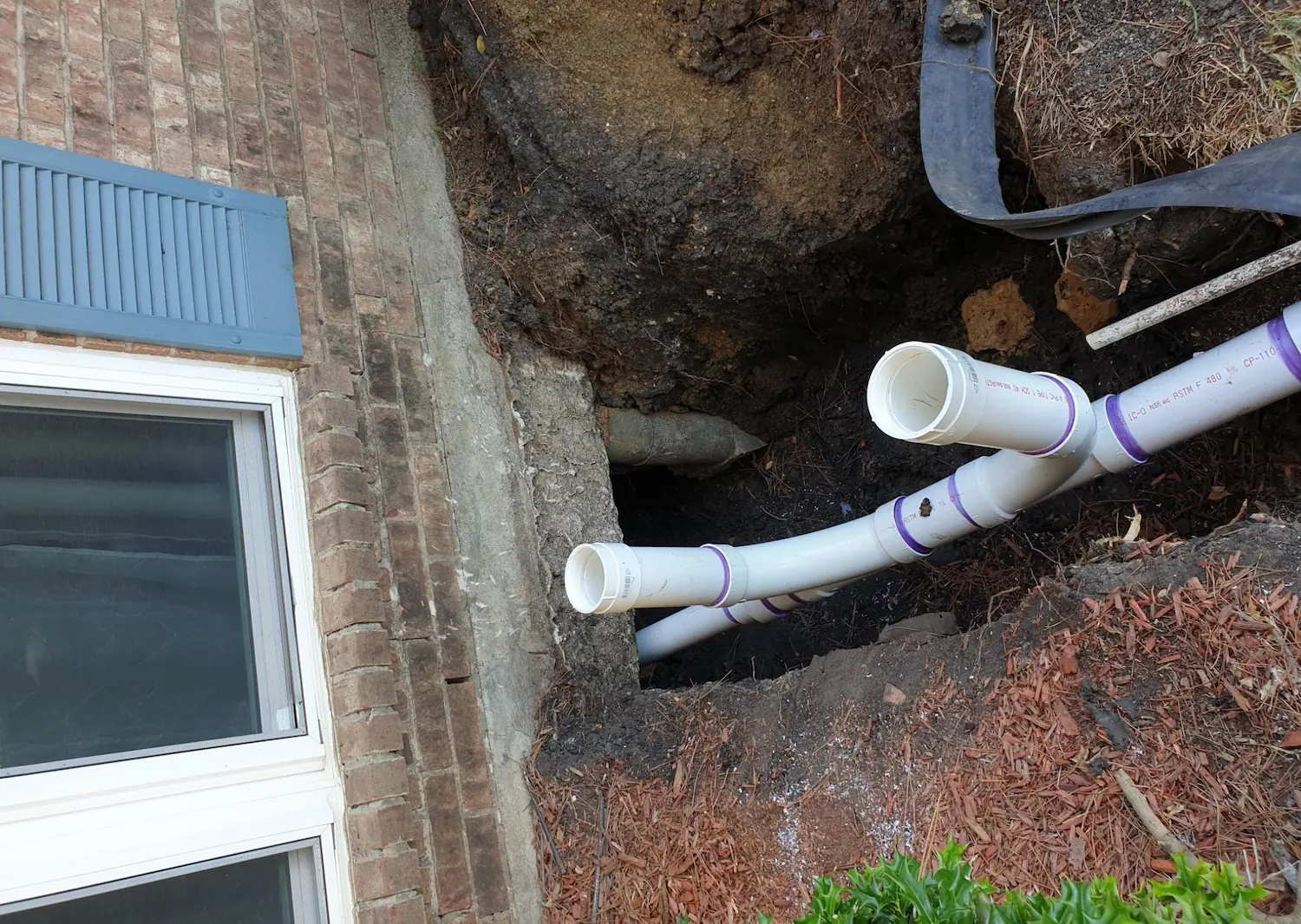 White PVC pipes being installed in a dug trench beside a brick house with a window.