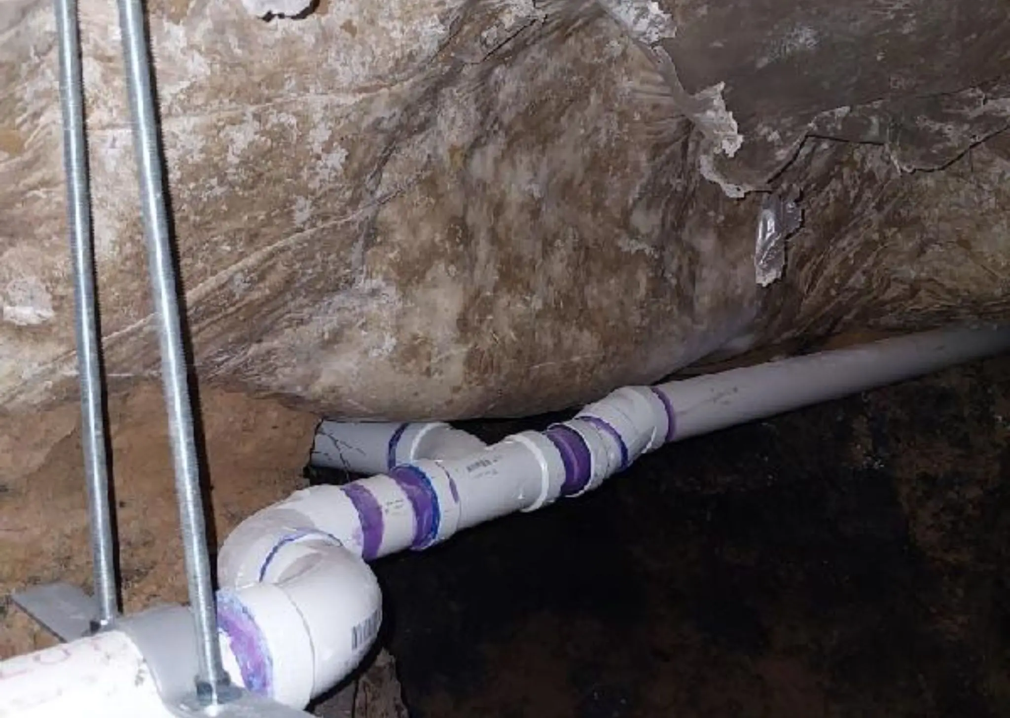 White PVC pipes with purple primer joints installed in a crawl space with a rocky ceiling.