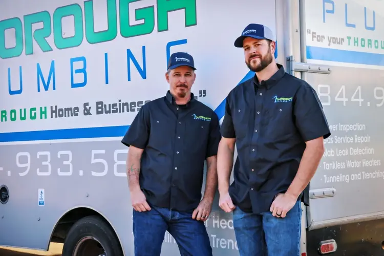 Two men in matching black plumber uniforms stand in front of a truck with plumbing company branding.
