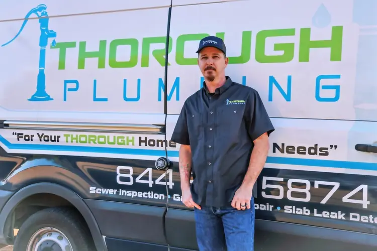 Man in plumbing uniform stands in front of a van with Thorough Plumbing logo and contact info.