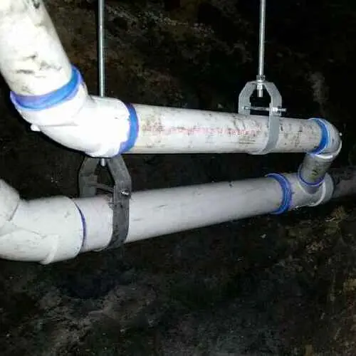 Two white PVC pipes with blue joints suspended by metal hangers in a dark space.