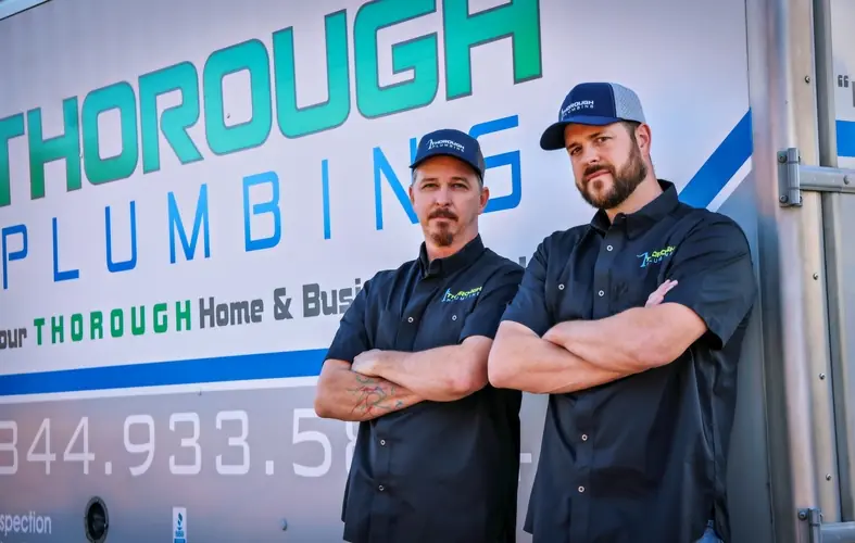 Two men in black shirts and caps with Thorough Plumbing logo standing with arms crossed by company truck.