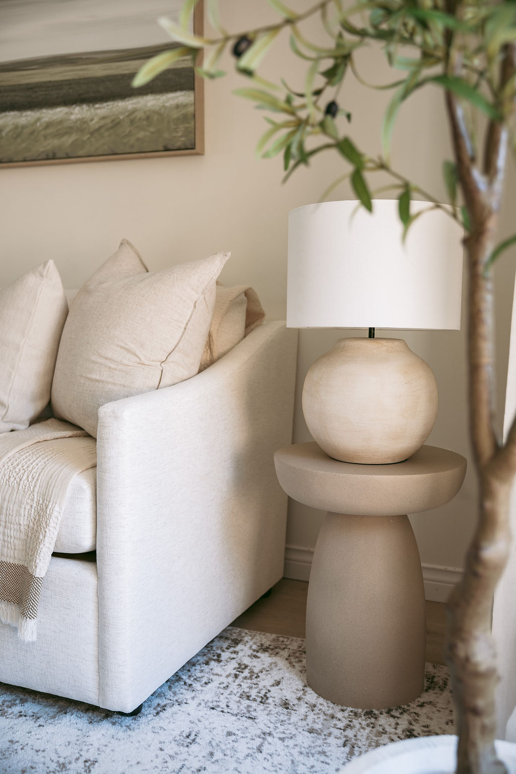  A brightly lit corner of a room features a modern white armchair with light wood accents, a small white coffee table with a plant, and a clear vase with white branches next to a sunlit window.