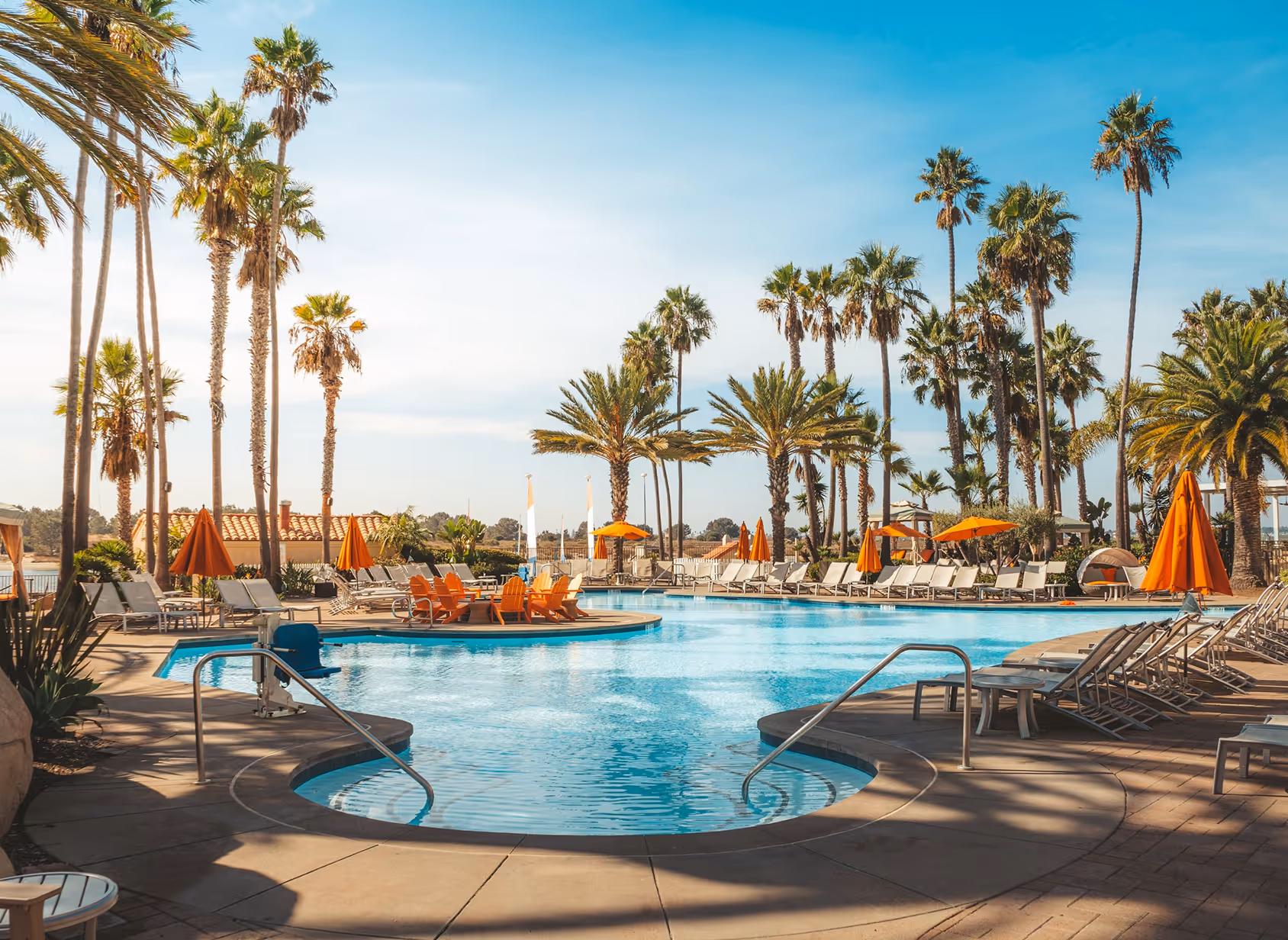 Clear outdoor pool with lounge chairs, orange umbrellas, and tall palm trees under blue sky.