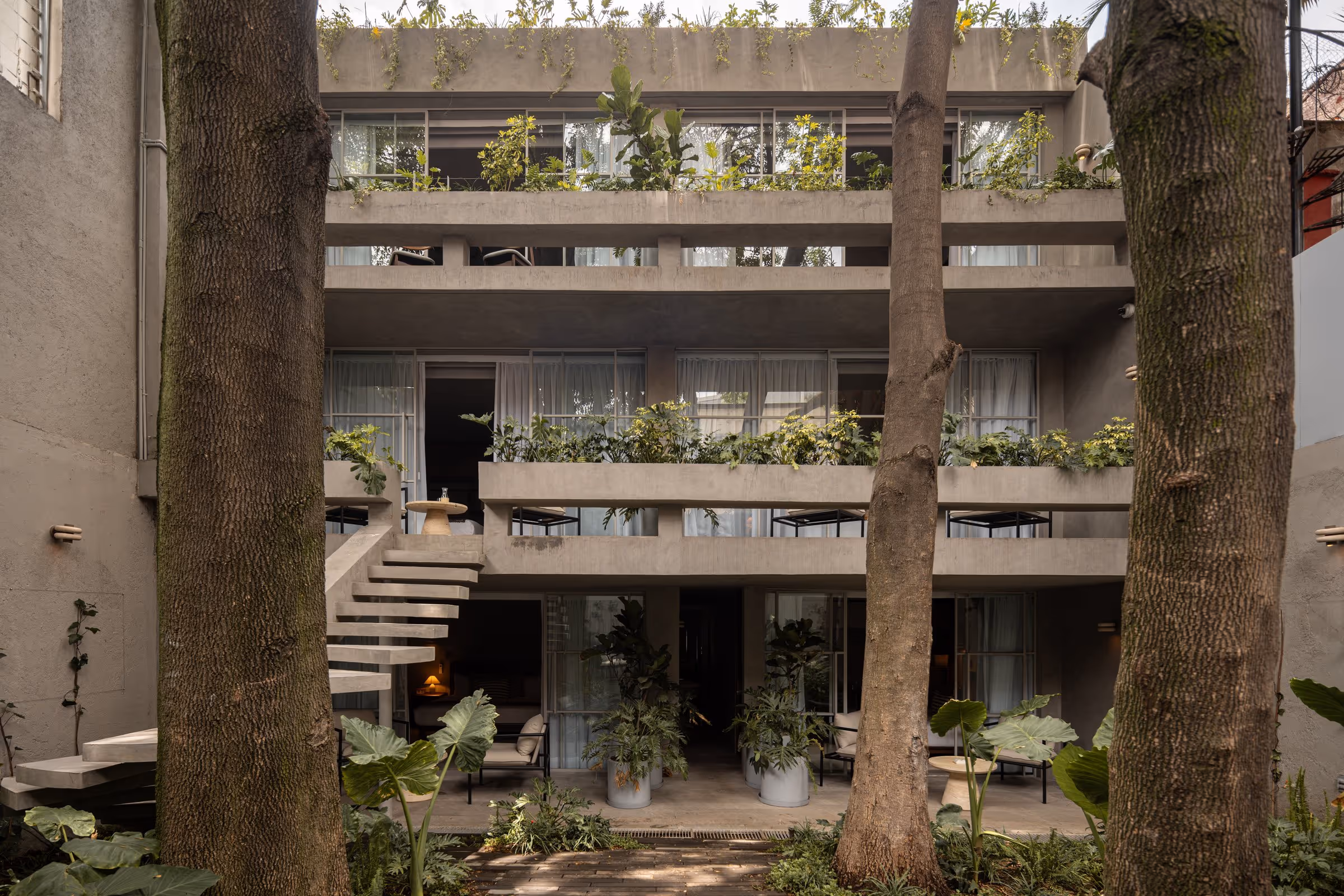 Modern three-story building with concrete balconies, plants, and large trees in the courtyard.