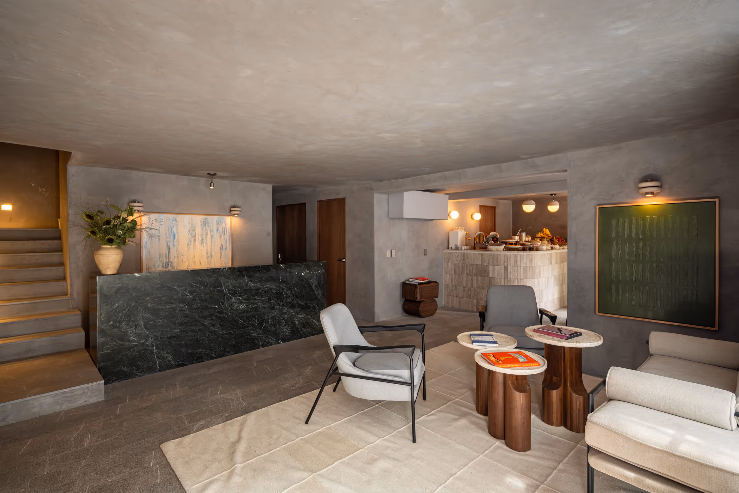 Modern lounge area with gray chairs, wooden tables with books, beige sofa, and a breakfast counter in the back.
