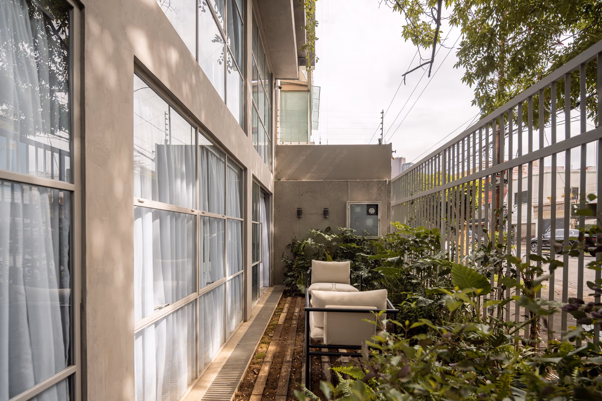 Outdoor seating area with two cushioned chairs among green plants beside a windowed building and metal fence.