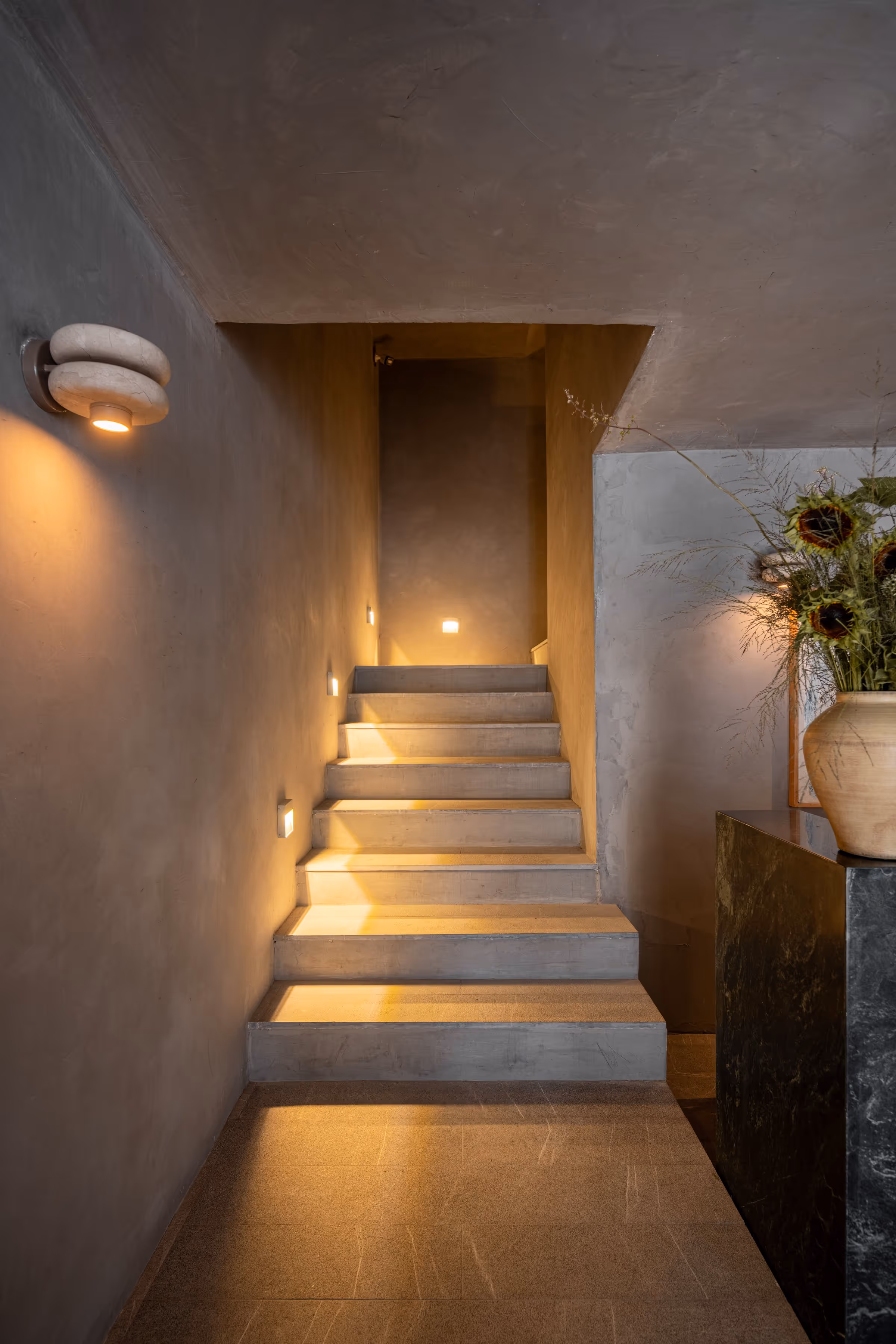 Minimalist concrete staircase lit by warm wall lights with a vase of sunflowers on a dark stand.