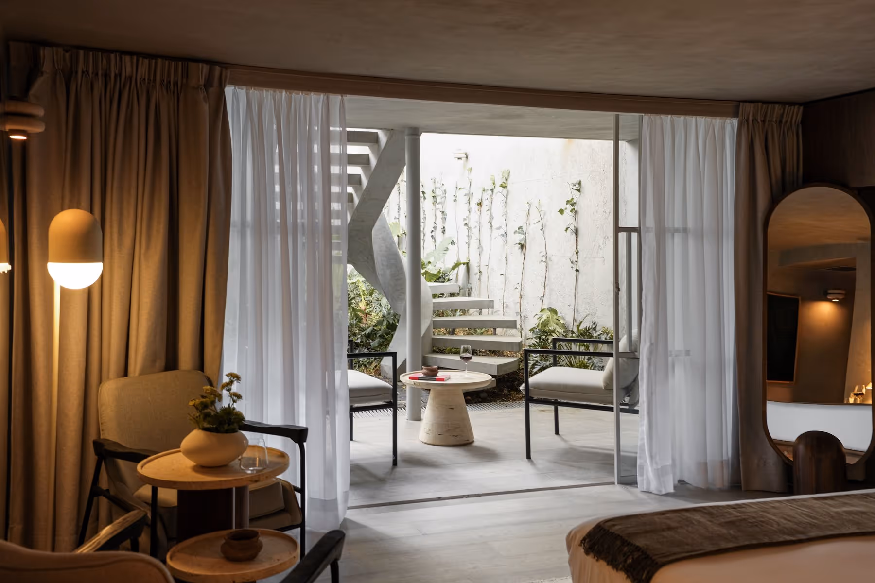Modern hotel room with neutral furniture, floor lamp, and patio with spiral stairs and seating.