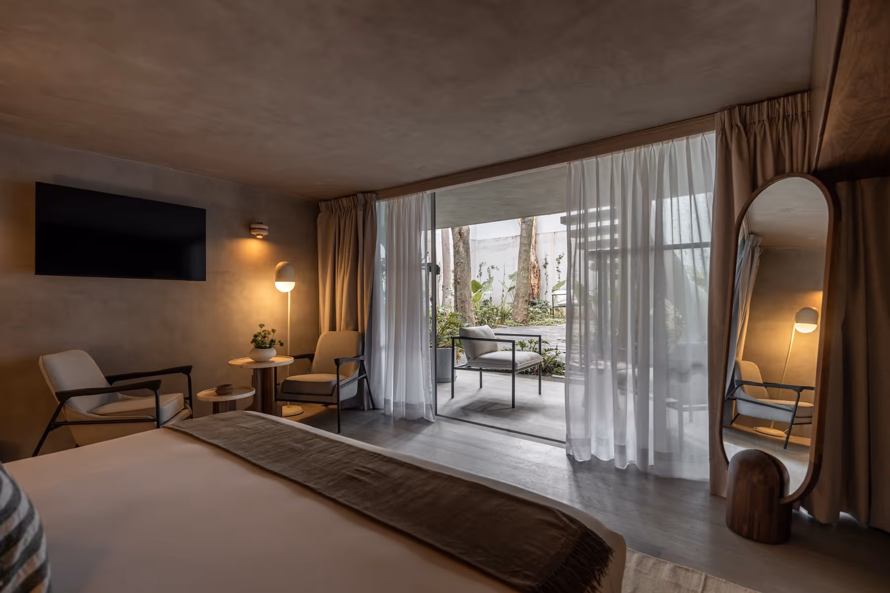 Modern bedroom with a bed, two armchairs, TV, floor lamp, and glass door to outdoor seating area.