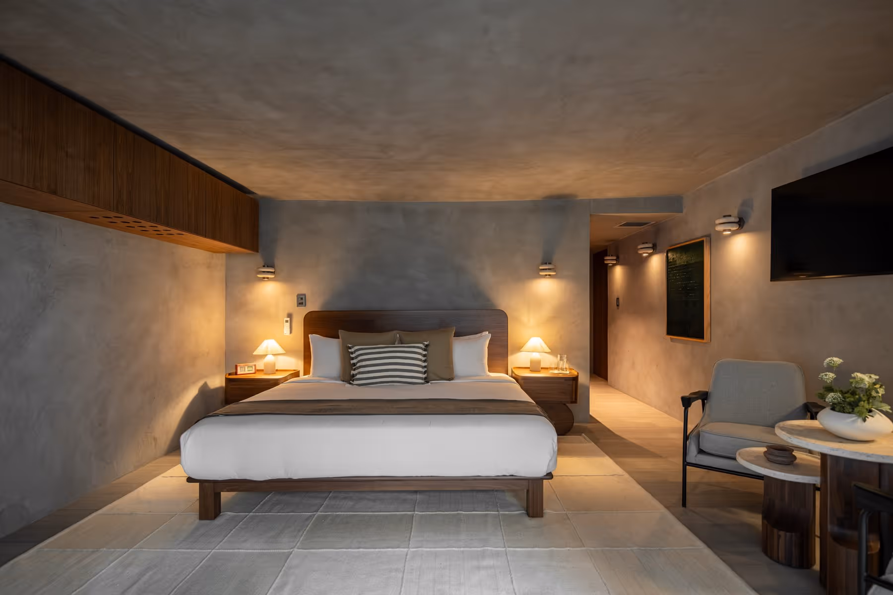 Modern bedroom with a king bed, two bedside lamps, gray walls, chair, round tables, and wall-mounted TV.