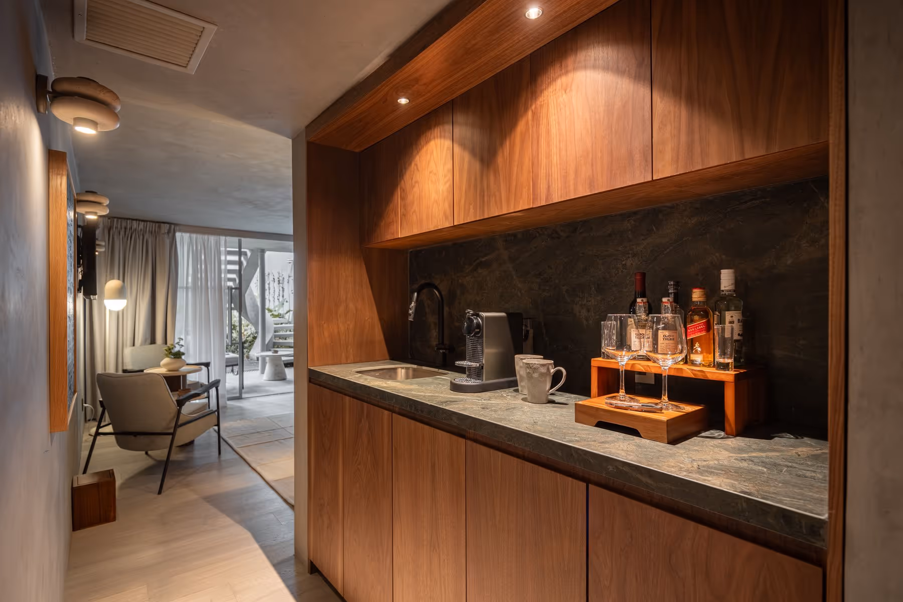 Compact kitchenette with wooden cabinets, stone countertop, coffee maker, mugs, and bottles under warm lighting.