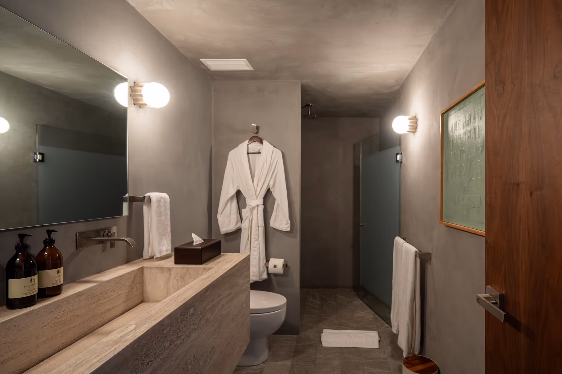 Modern bathroom with stone sink, toilet, white robe, glass shower, towel racks, and wooden door.