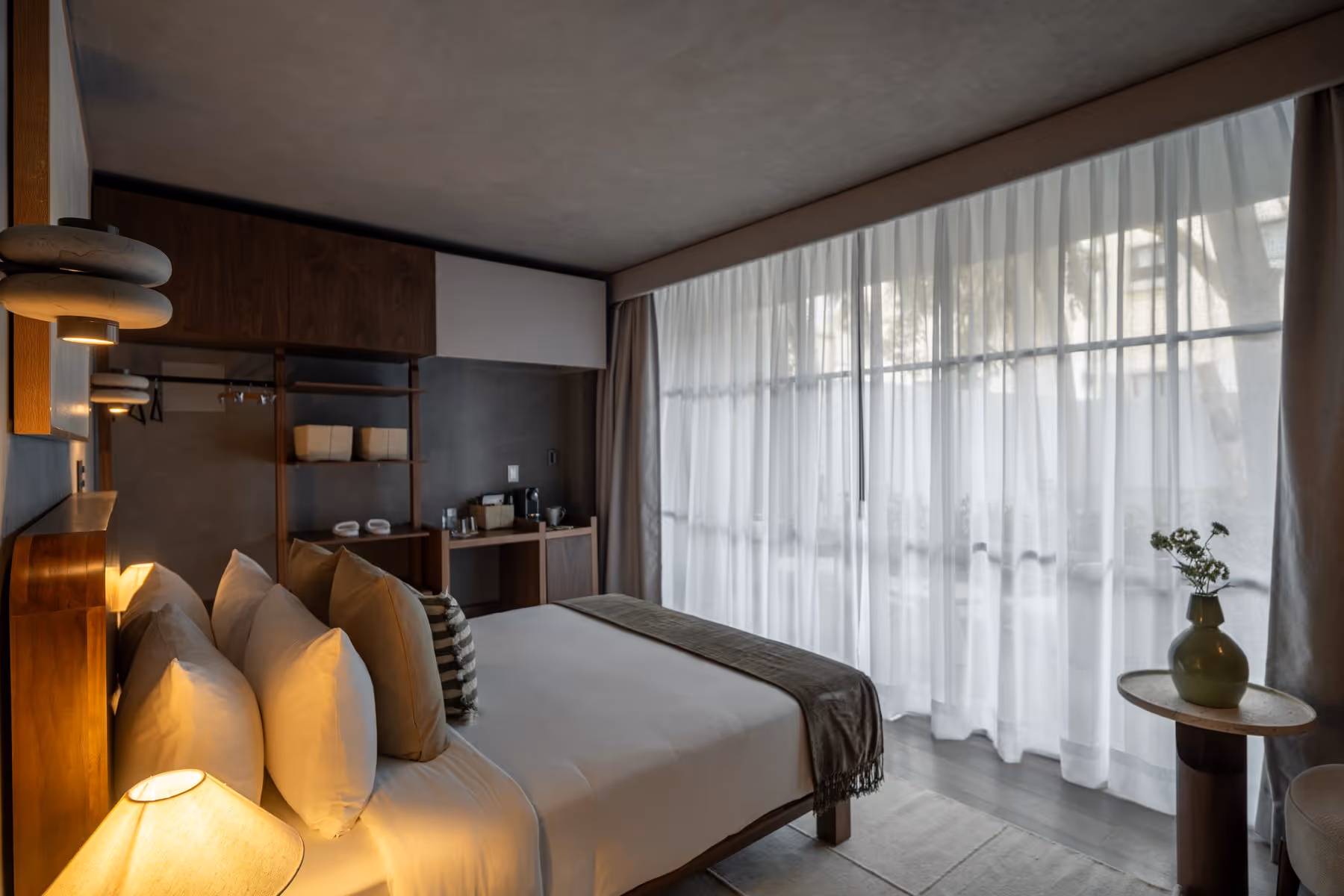 Modern bedroom with a large bed, multiple pillows, wooden furniture, and sheer curtains over a window.
