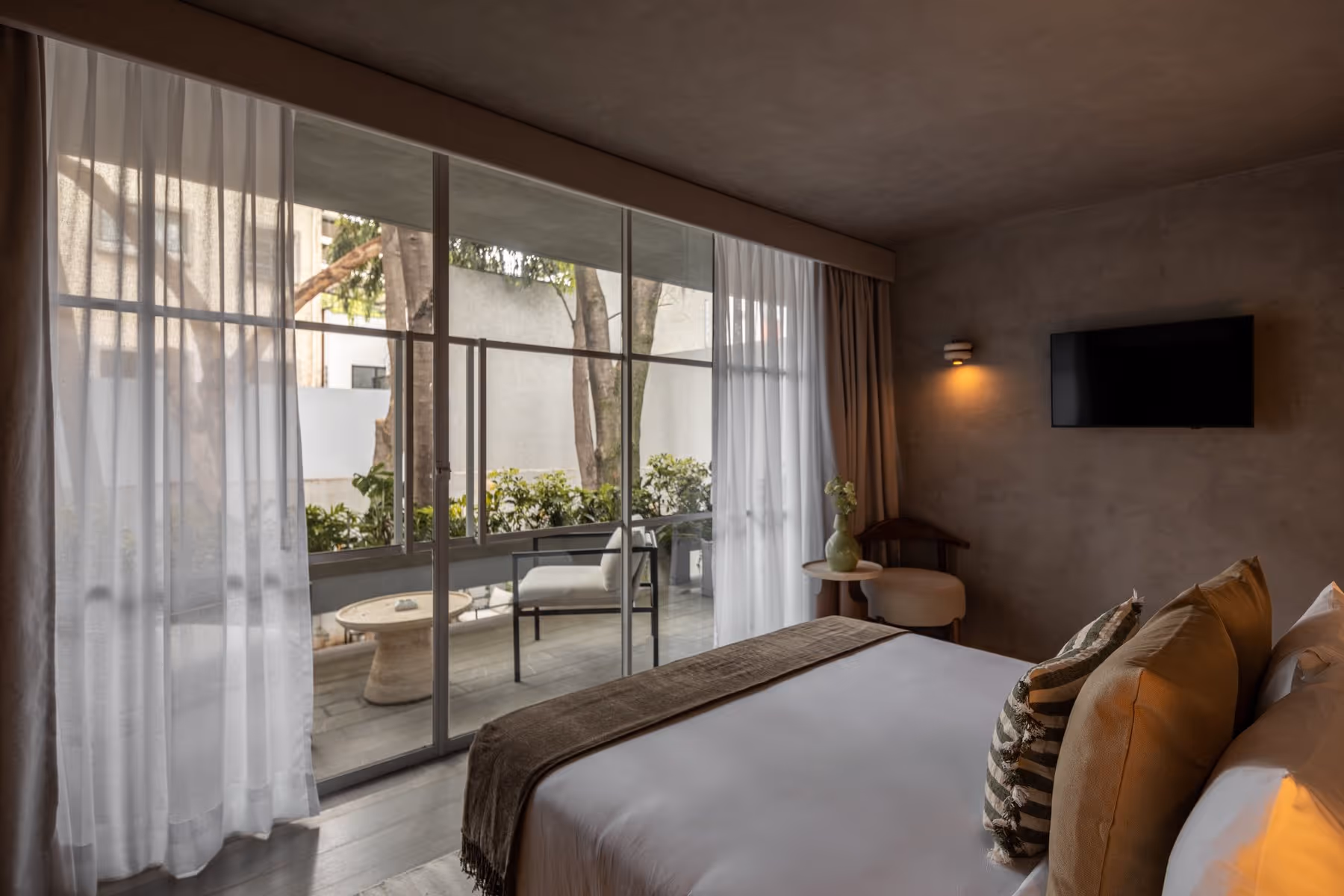 Cozy hotel room with queen bed, pillows, side chair, round table, TV, and terrace with outdoor seating.