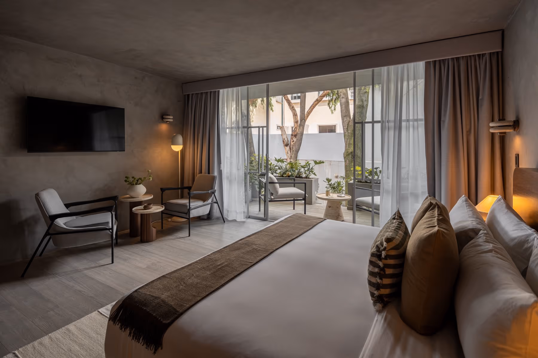 Modern hotel room with bed, pillows, two armchairs, a TV, and sliding glass door to patio with plants.