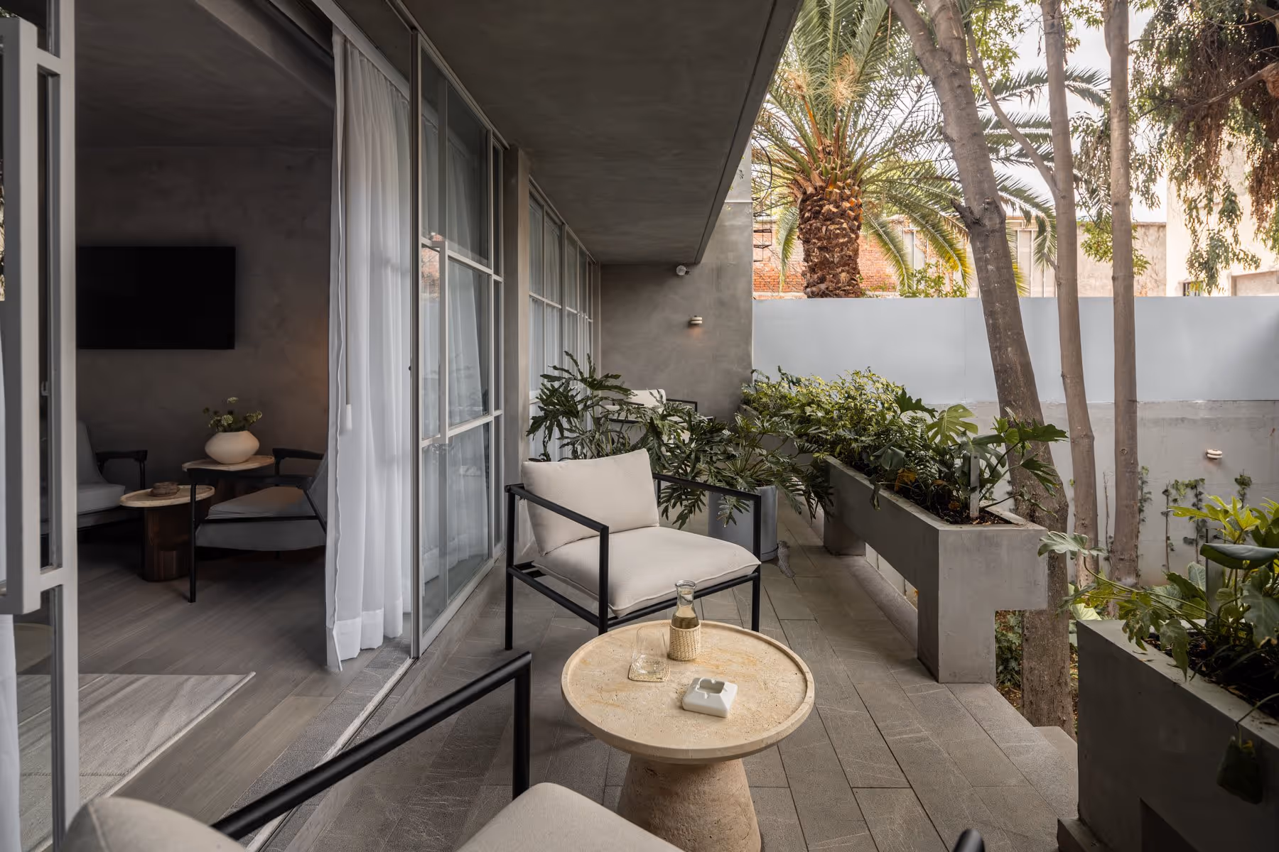 Modern terrace with cushioned chairs, a stone table with a carafe, surrounded by potted plants and trees.