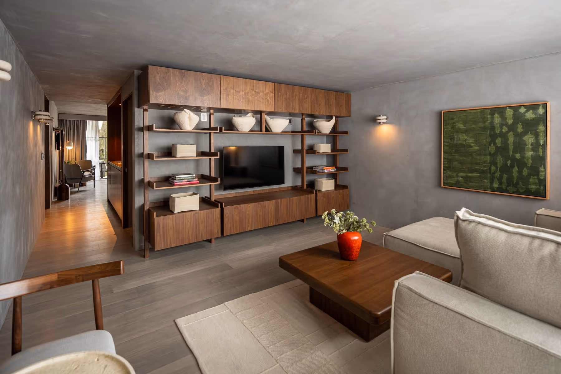 Modern living room with wooden shelving, TV, beige sofa, coffee table with red vase, and green abstract wall art.