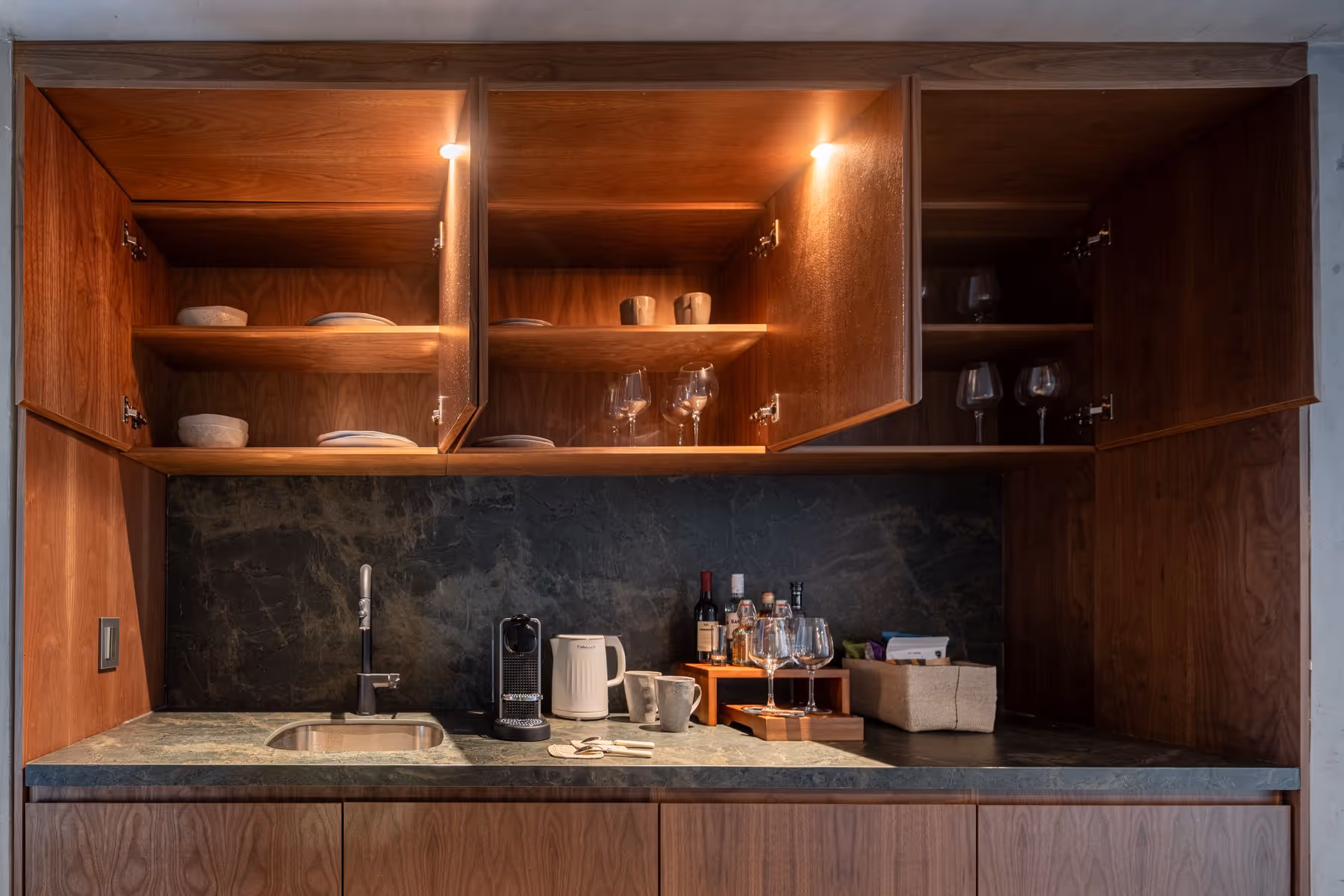 Modern kitchenette with open wooden cabinets, sink, coffee machine, kettle, cups, and wine glasses.