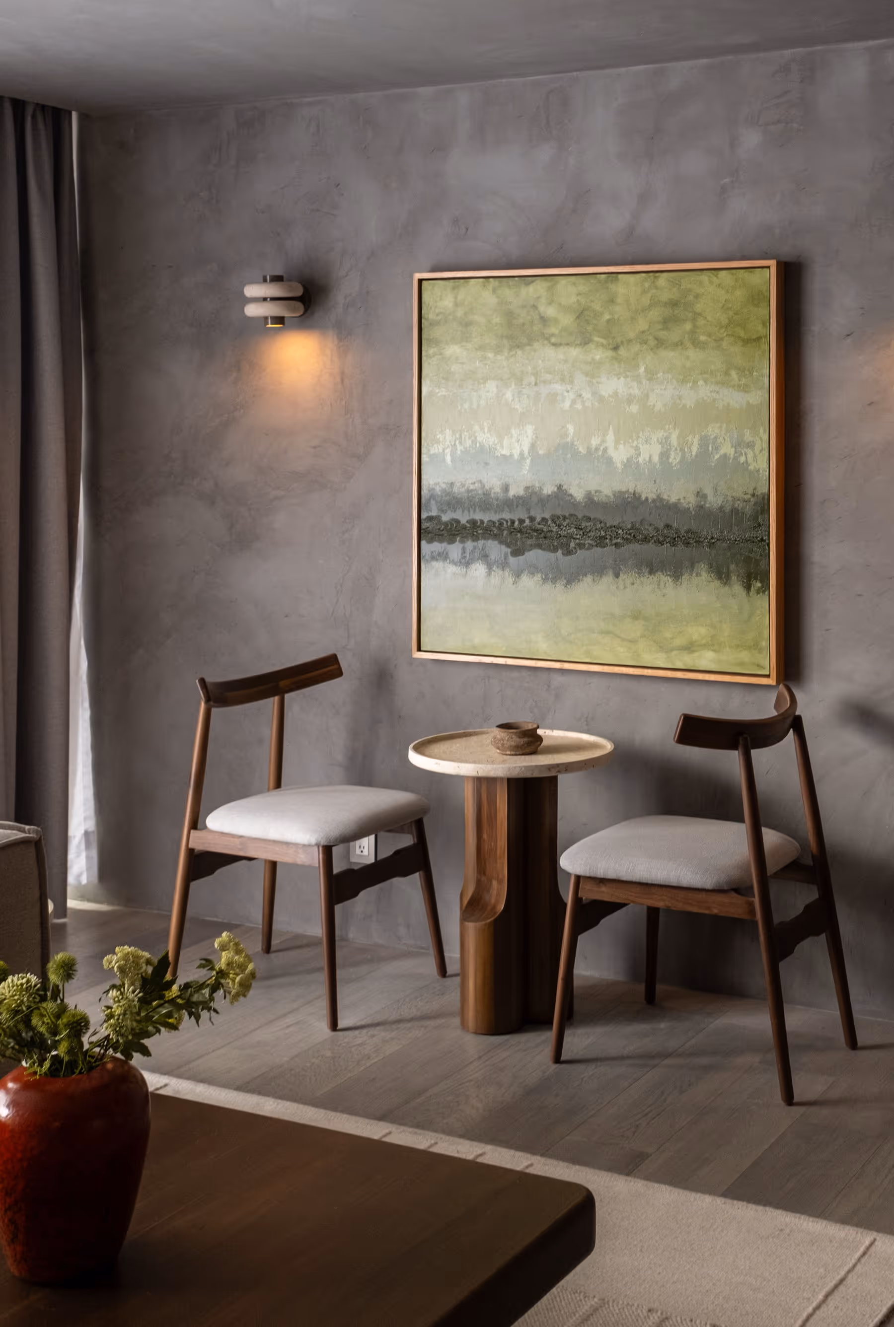 Modern sitting area with two wooden chairs, a round table, green wall art, and a vase with flowers.
