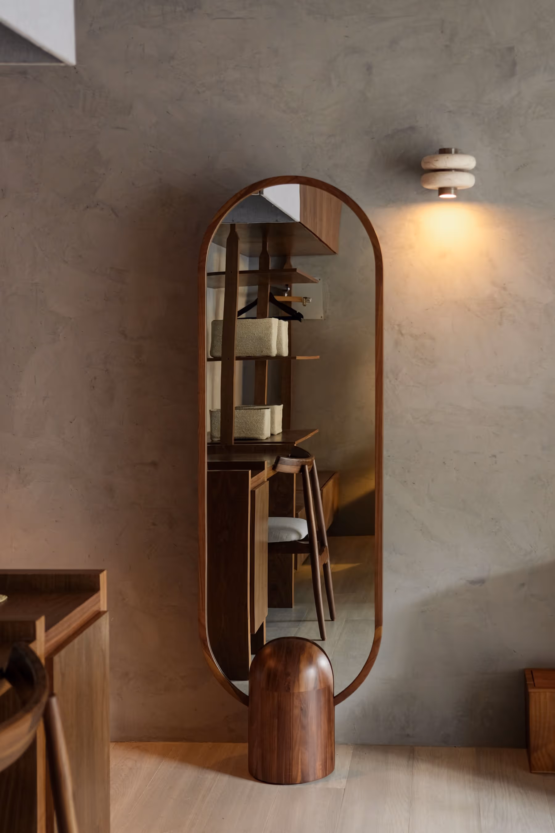 Tall wooden-framed mirror reflects a chair and shelves in a room with textured gray walls and soft lighting.