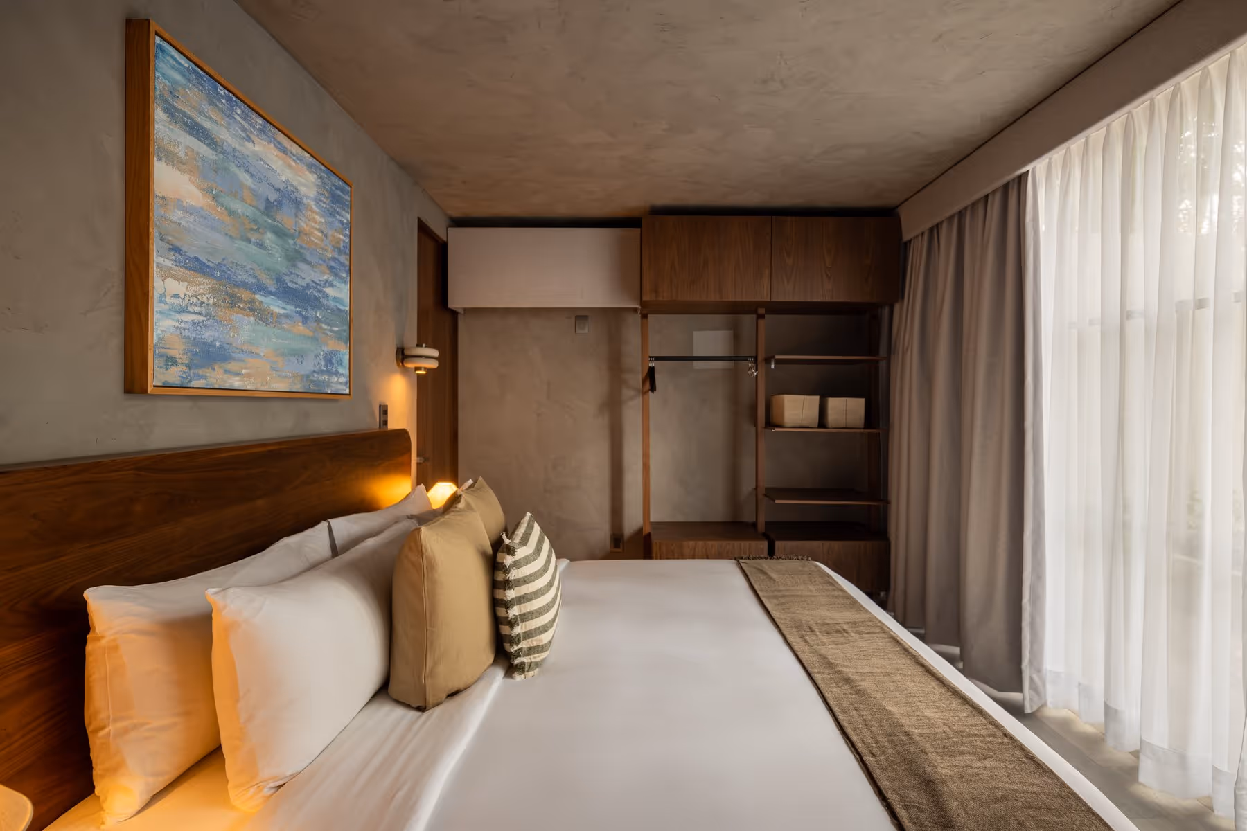 King-size bed with white linens and pillows, abstract wall art, wooden shelves, and sheer curtains in room.