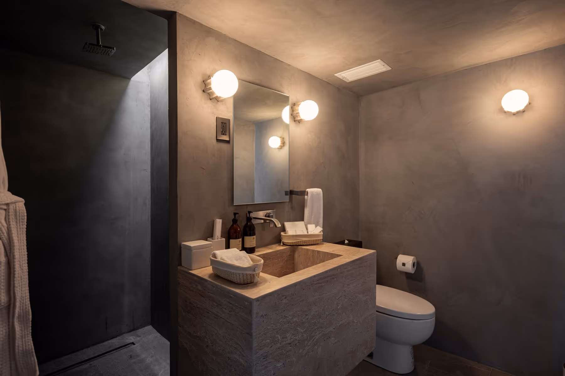 Modern bathroom with stone sink, wall mirror, two lights, toilet, and dark shower area.