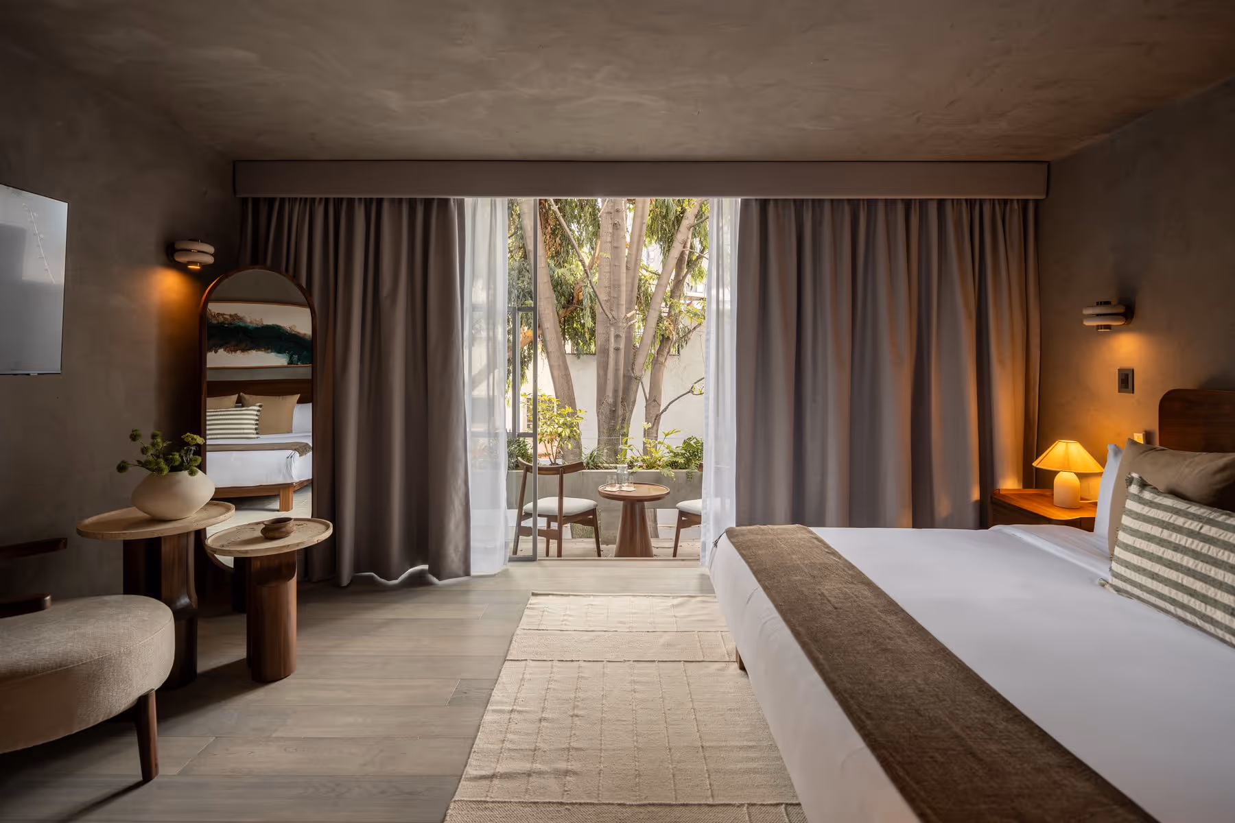 Modern bedroom with king-size bed, bedside lamps, mirror, and outdoor seating visible through glass doors.