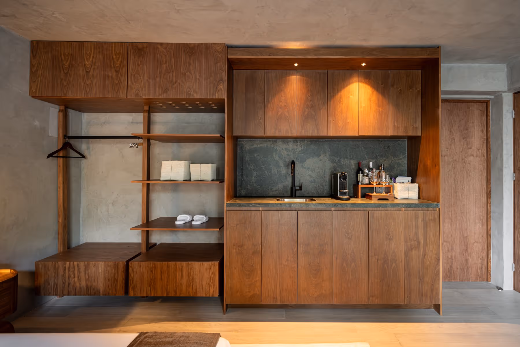 Modern kitchenette with wooden cabinets, open shelving, sink, coffee maker, and assorted bottles and glasses.