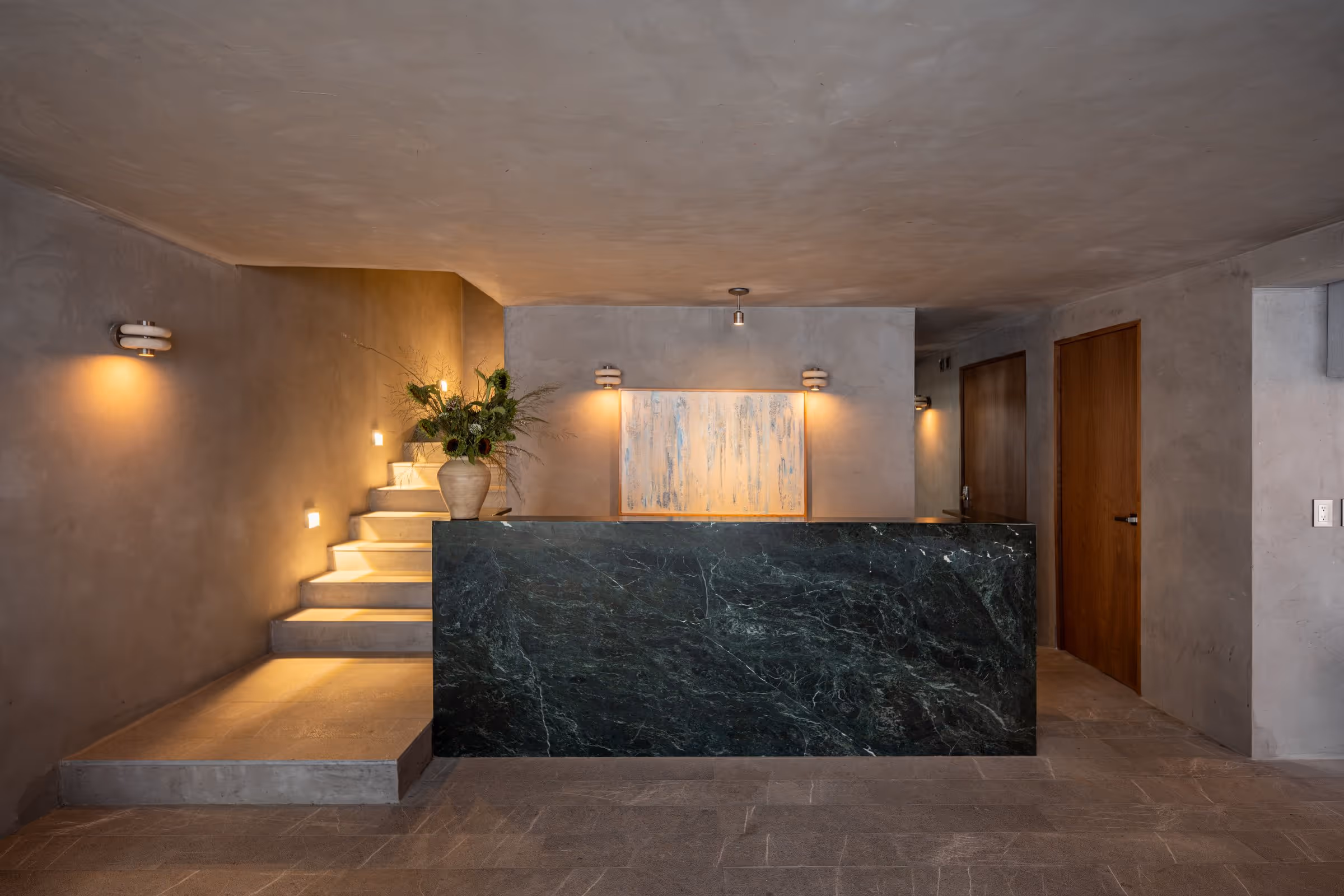 Modern lobby with dark green marble reception desk, concrete walls, stairs, and wall-mounted lights.