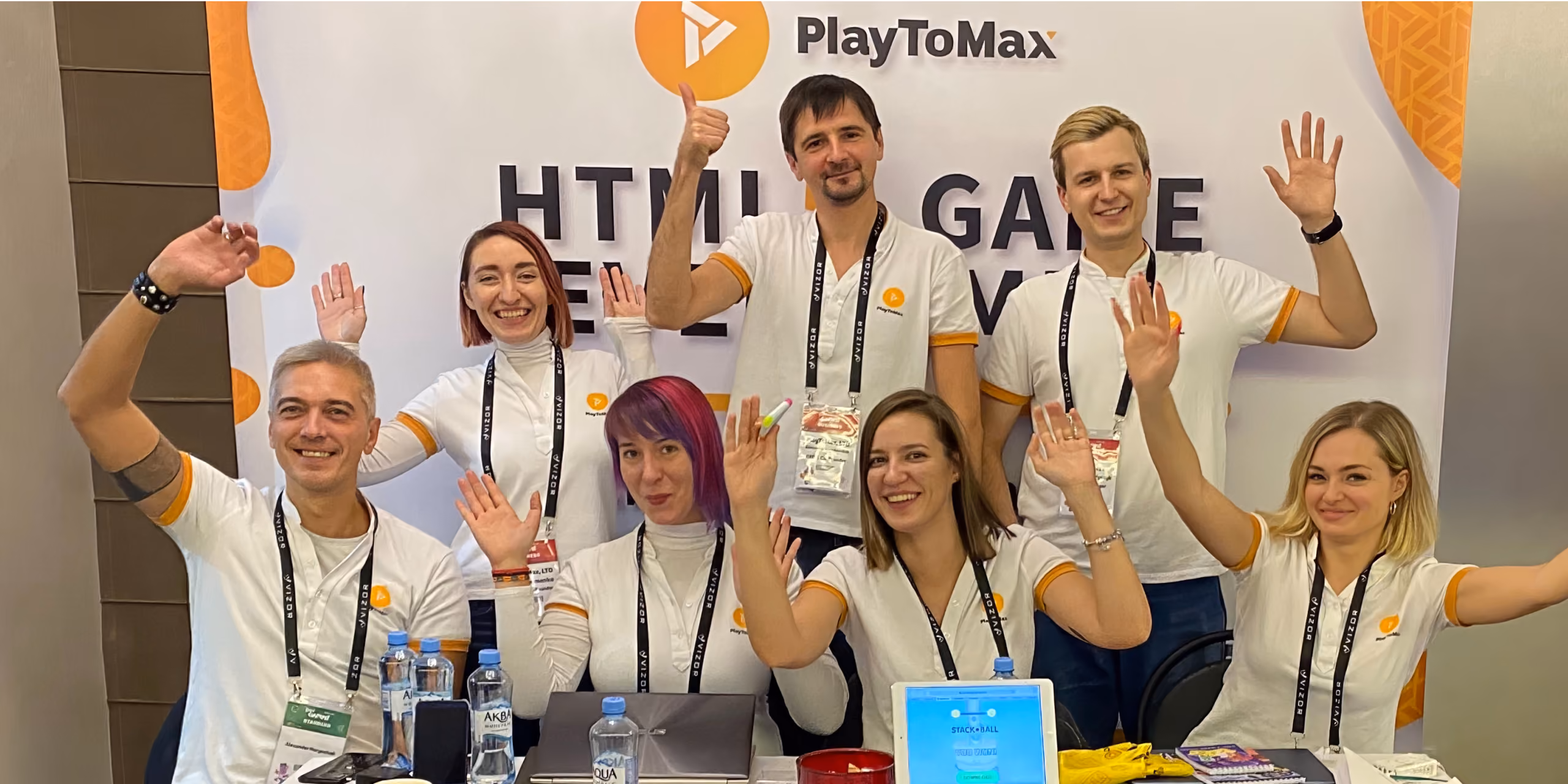 Seven smiling team members in white PlayToMax shirts waving at a conference booth.
