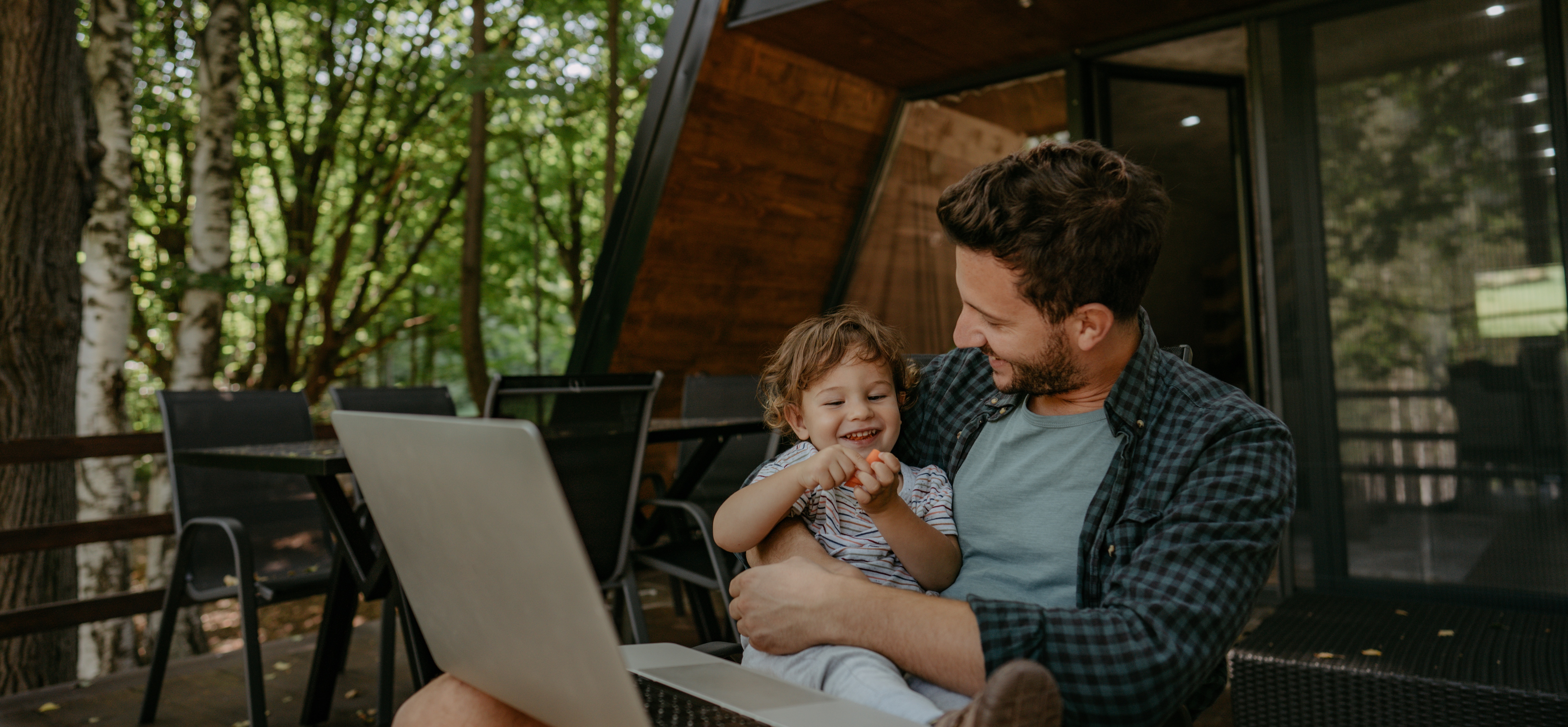 Man holding smiling young child on patio with laptop, surrounded by outdoor greenery and wooden house.