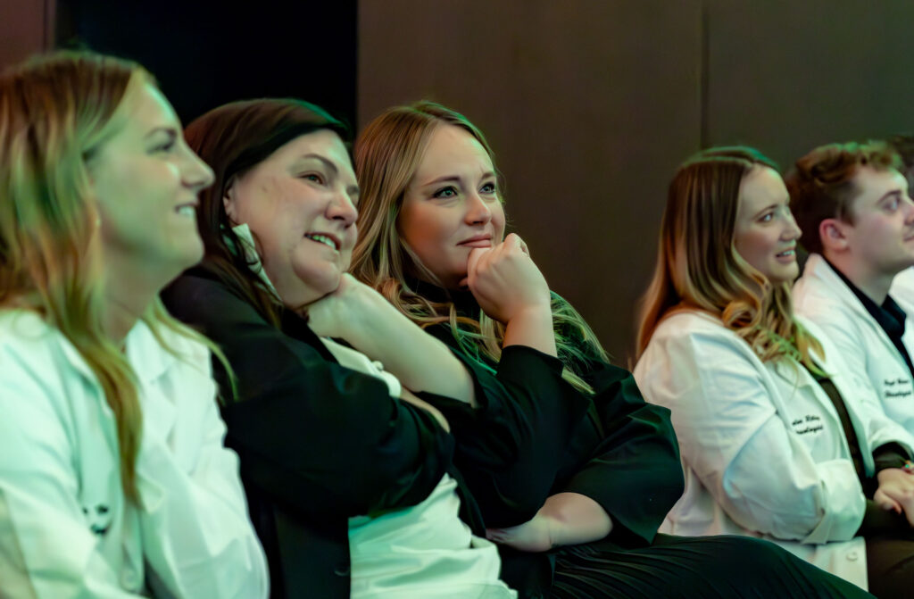 Group of young adults wearing lab coats attentively watching something off camera.