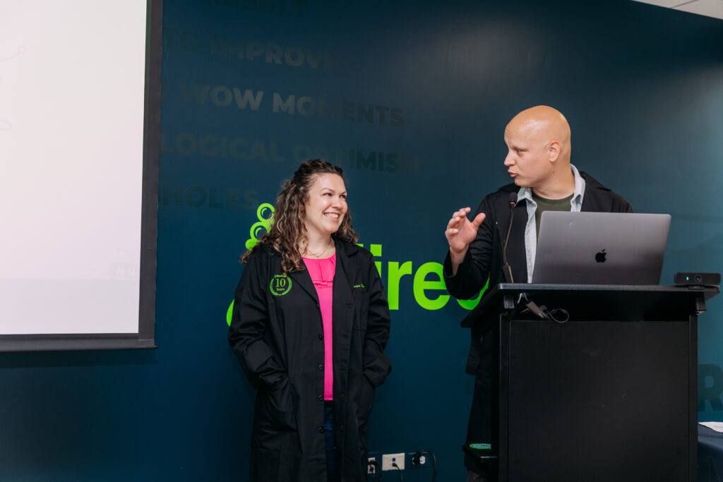 A woman smiling and a man speaking at a podium with a laptop in front of a dark blue wall.