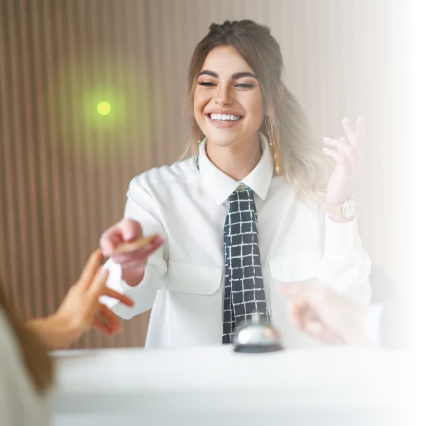 Smiling woman in white shirt and patterned tie hands an item to a guest at a reception desk.
