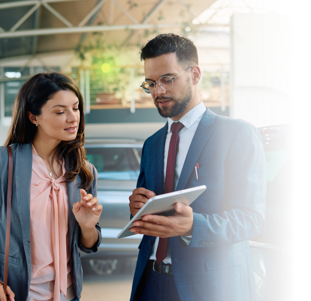Man in suit with glasses shows tablet to woman as they discuss inside a car showroom.