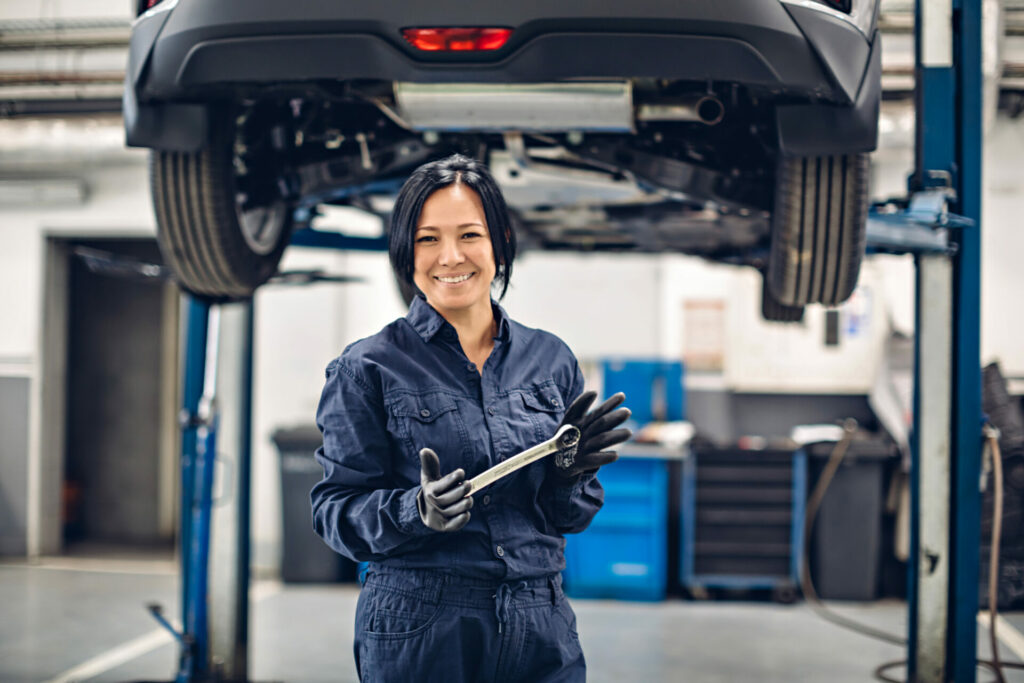 Smiling female mechanic in blue coveralls holding wrench under car on lift in workshop.