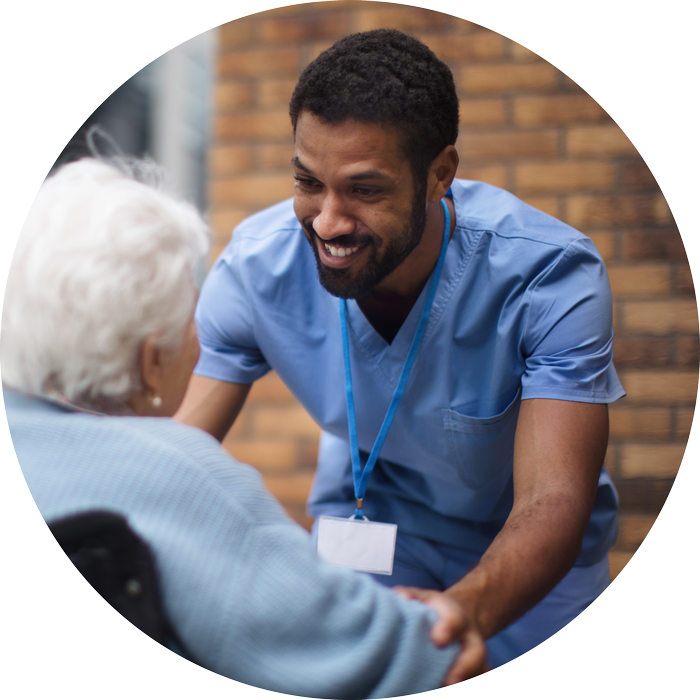 Smiling healthcare worker in blue scrubs warmly holding hands of an elderly person in a wheelchair.