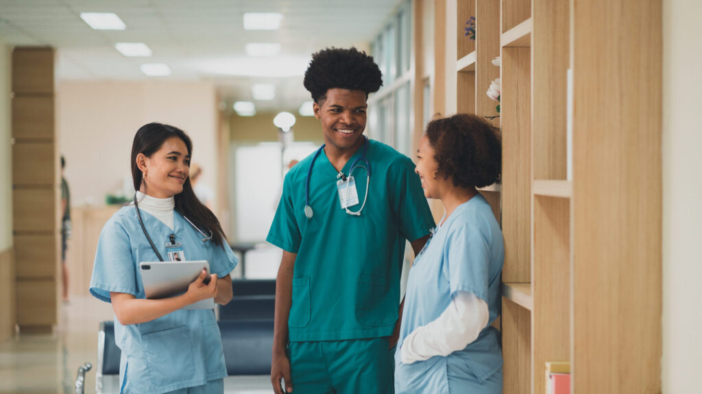 Three medical professionals in scrubs chatting and smiling in a hospital hallway.
