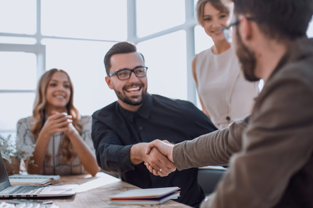 Two men shaking hands across a table with two smiling women nearby in a bright office setting.