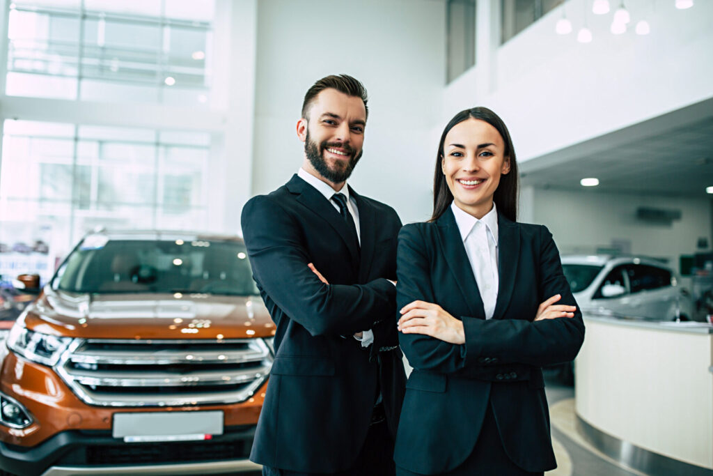 Man and woman in business suits smiling with crossed arms in a car dealership showroom.