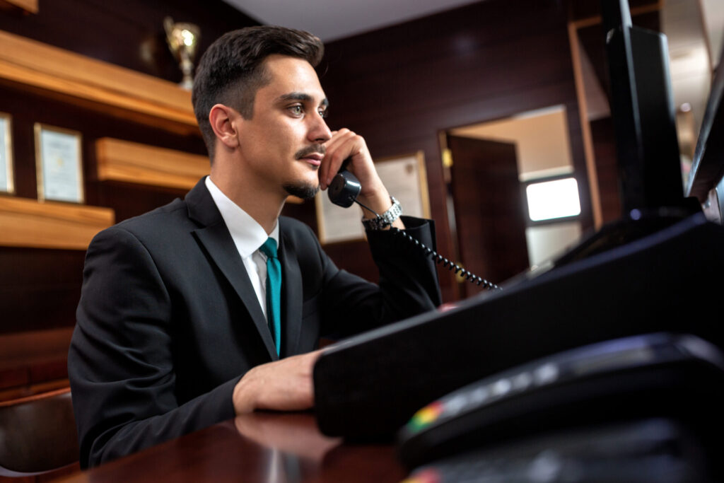 Man in suit talking on a corded telephone at an office desk with computer monitors.